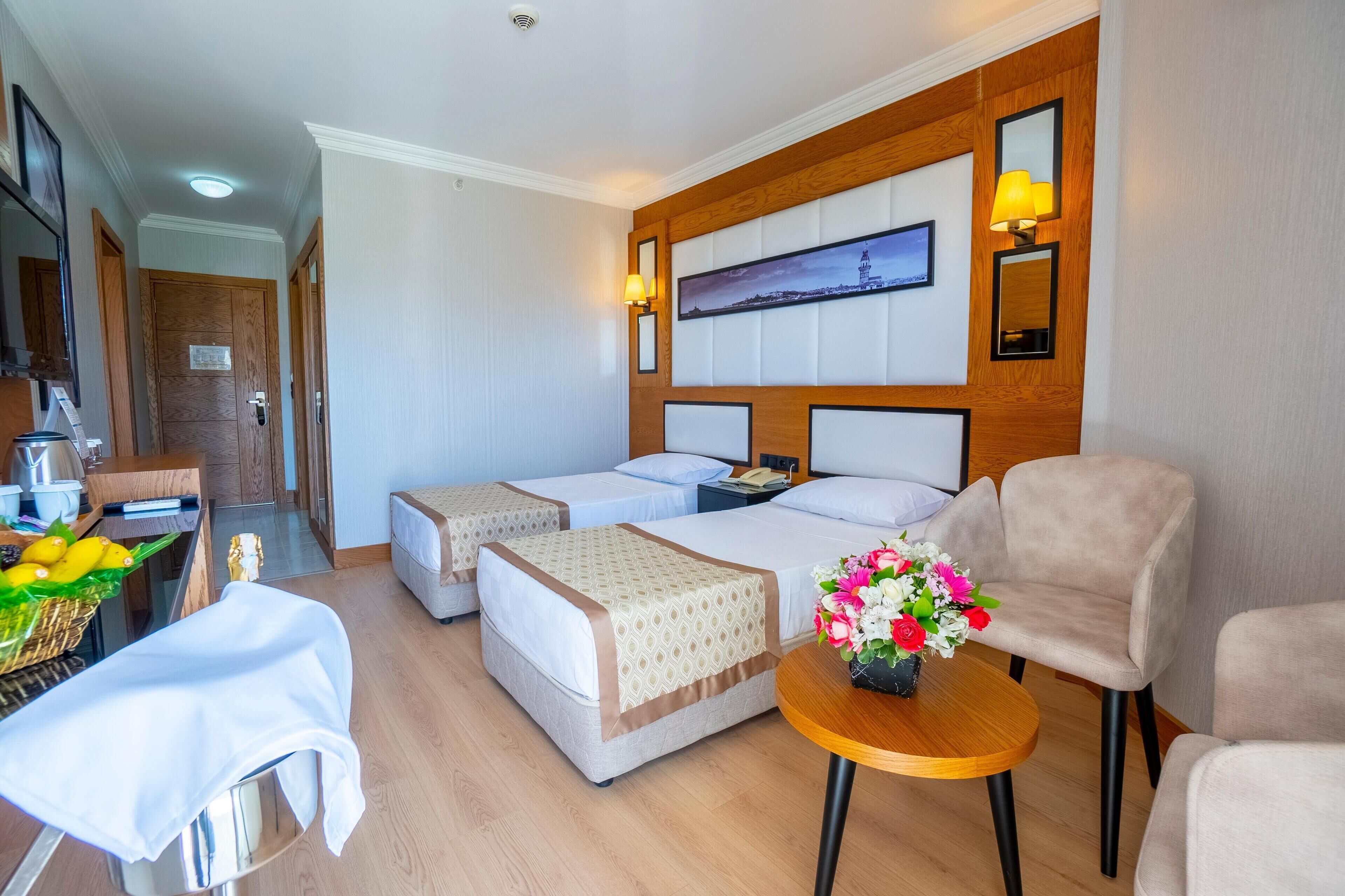 Une chambre d’hôtel avec deux lits, une petite table ornée d’un vase de fleurs, une chaise et un bureau avec un panier de fruits.