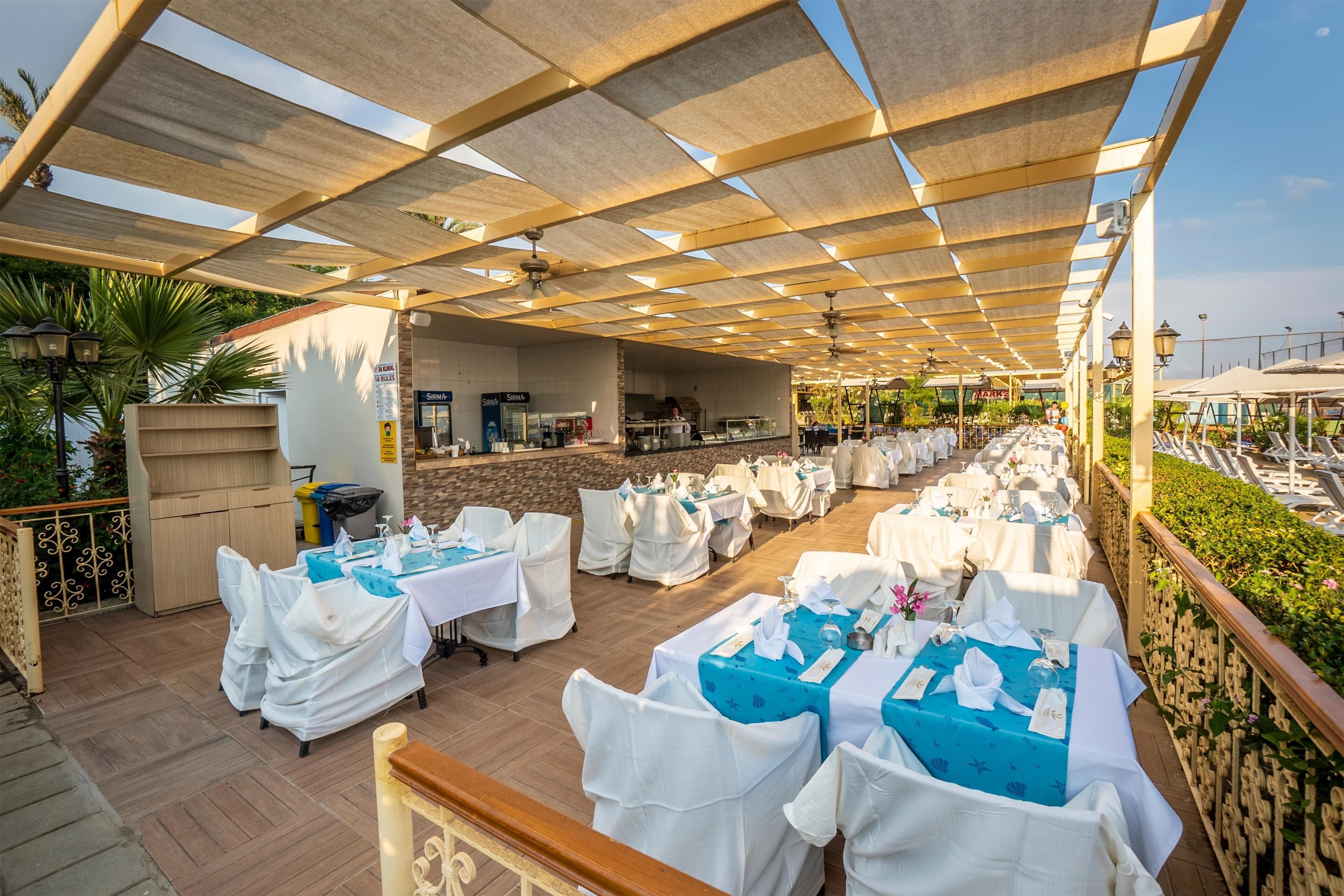 An outdoor dining area with tables covered in white and blue tablecloths, a kitchen area, and a view of the sea.