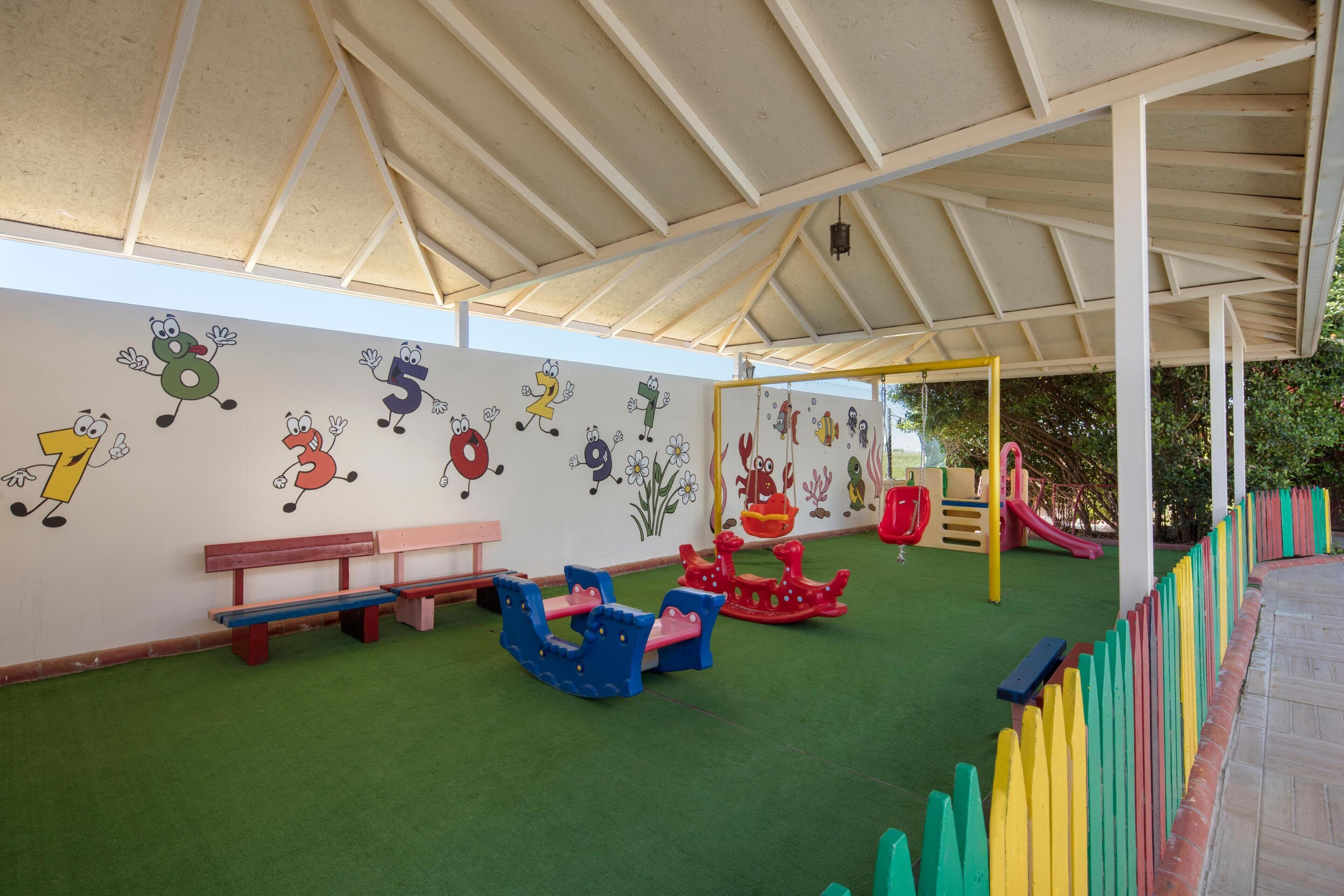 A children's play area with a covered roof, benches, and colorful play equipment.