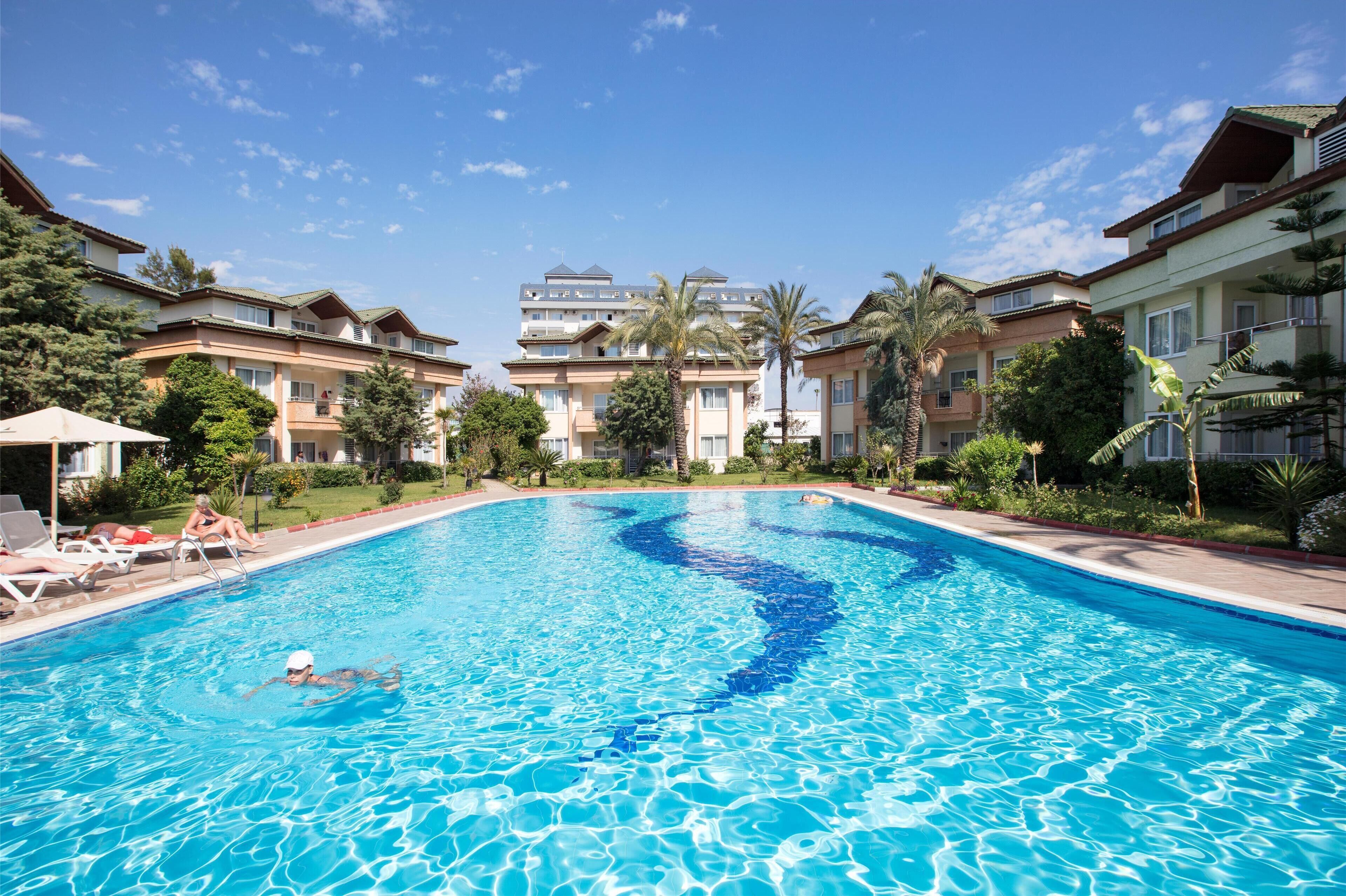 A swimming pool with clear blue water, surrounded by multi-story buildings with balconies and palm trees.