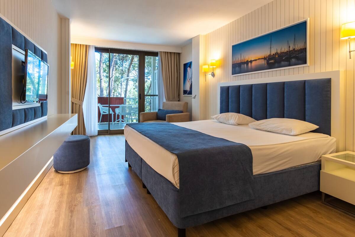 Habitación de hotel moderna con una cama grande, televisor, balcón con mesa y sillas, y una pintura de paisaje urbano.
