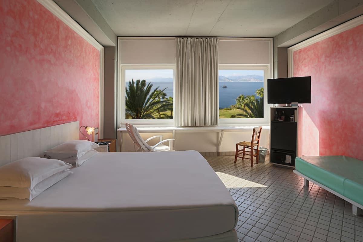 A hotel room with a large bed, a desk with a lamp, a chair, a TV, and a window with a view of the sea and mountains.