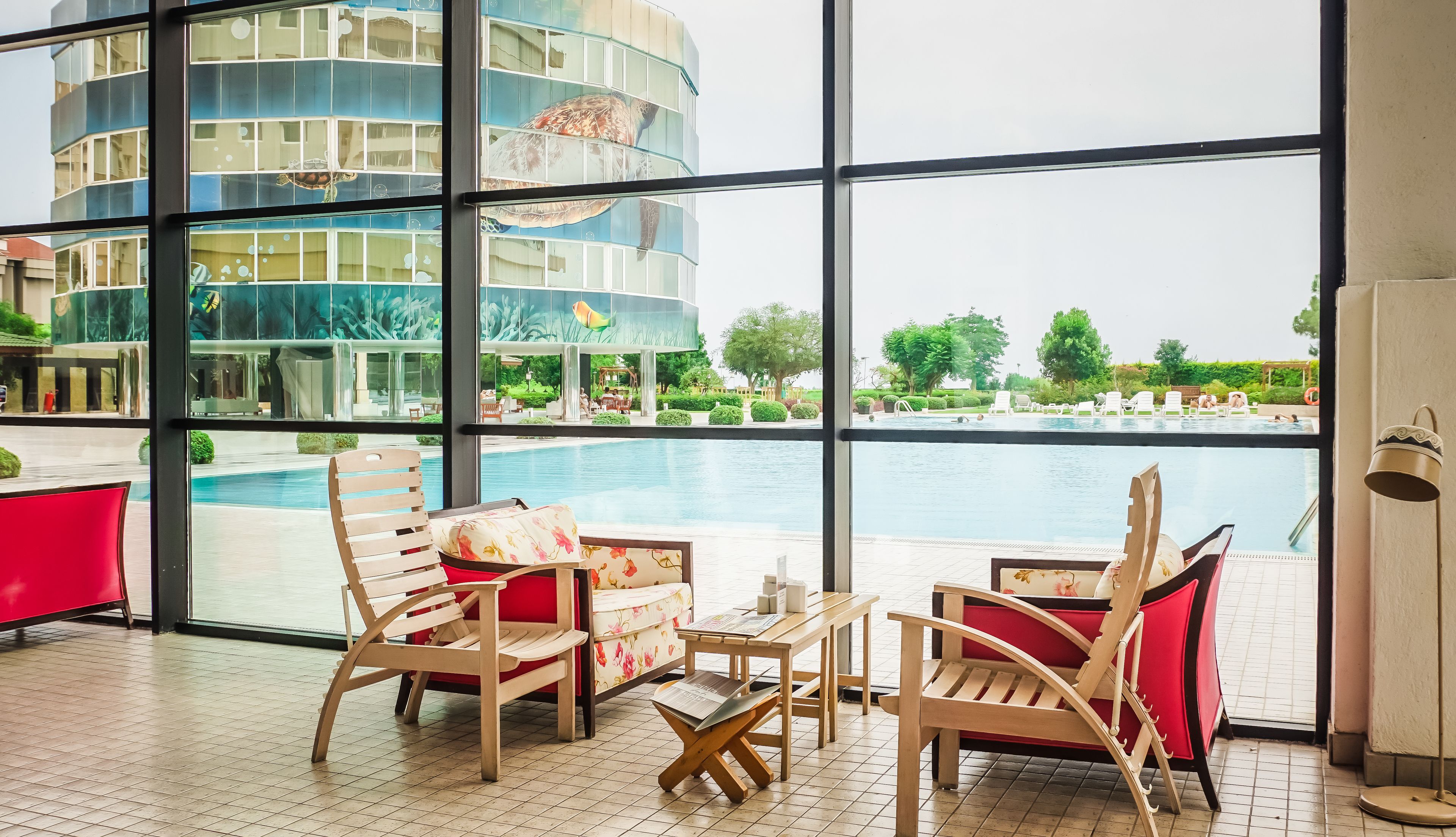 A modern hotel lobby with large windows overlooking a swimming pool and a landscaped garden.