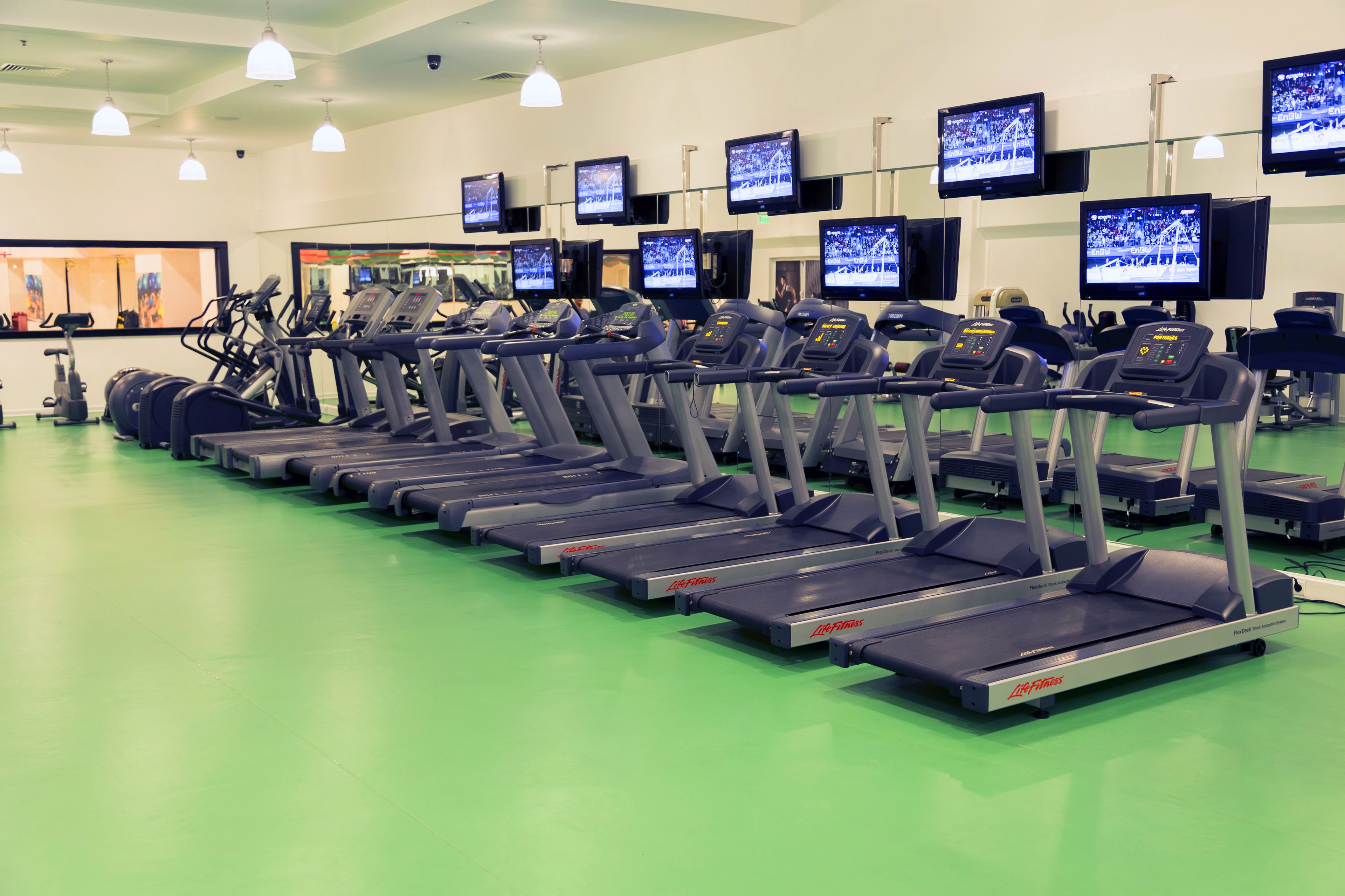 A gym with multiple treadmills aligned in rows, each with a control panel and a screen displaying a running event.