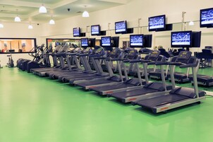 A gym with multiple treadmills aligned in rows, each with a control panel and a screen displaying a running event.