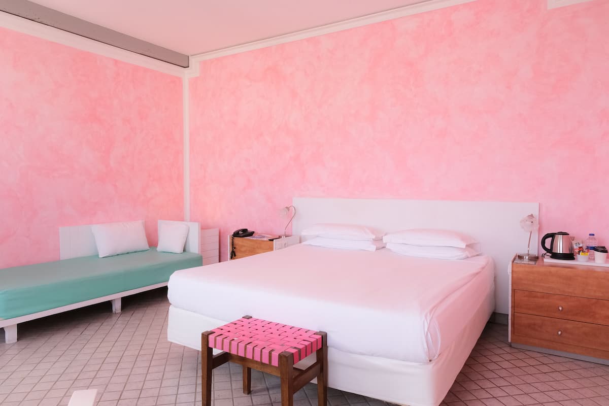 A bedroom with a large bed, a smaller bed, a wooden nightstand, a wooden dresser, and a pink woven stool.
