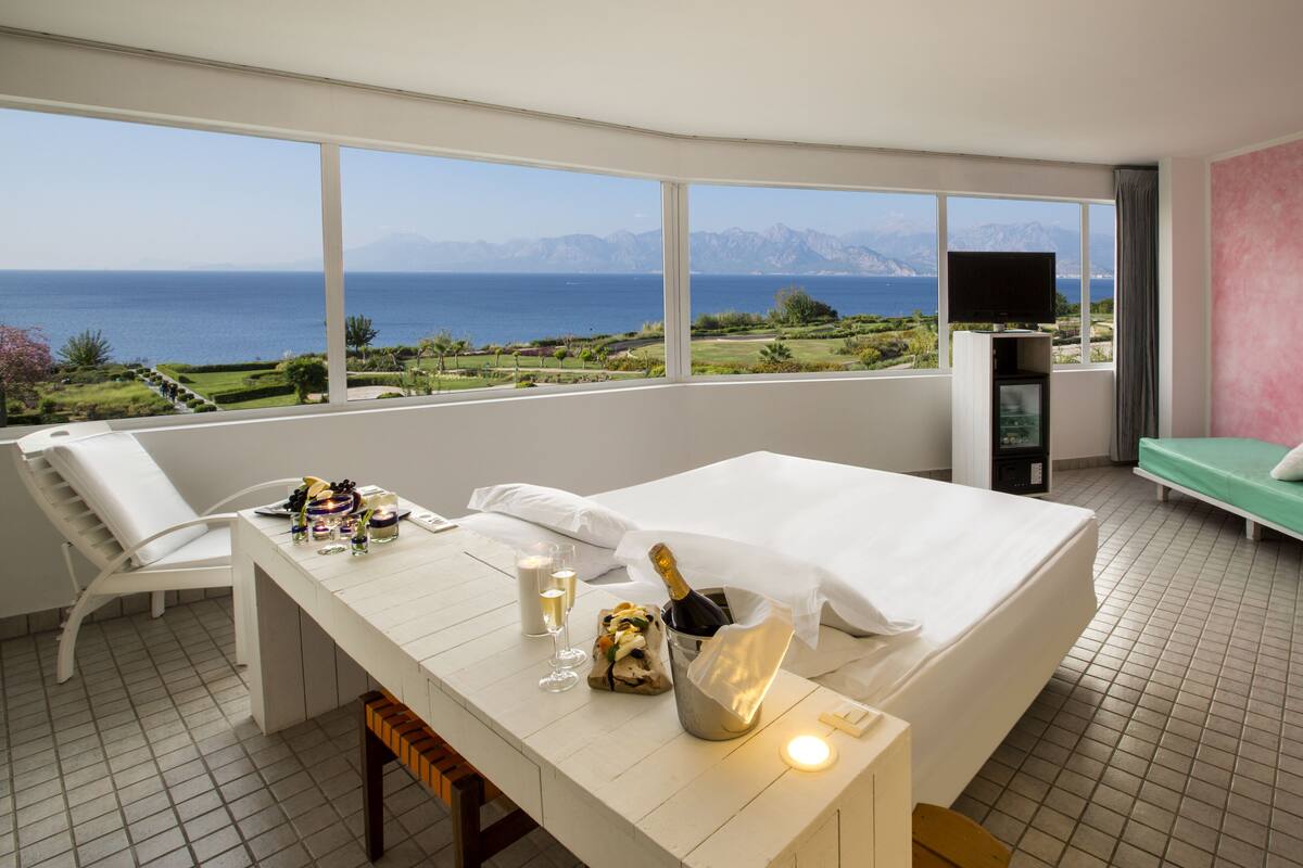 A hotel room with a large window offering a scenic view of the sea and mountains. There is a bed, a small table with a bottle of champagne, and a TV on a stand.