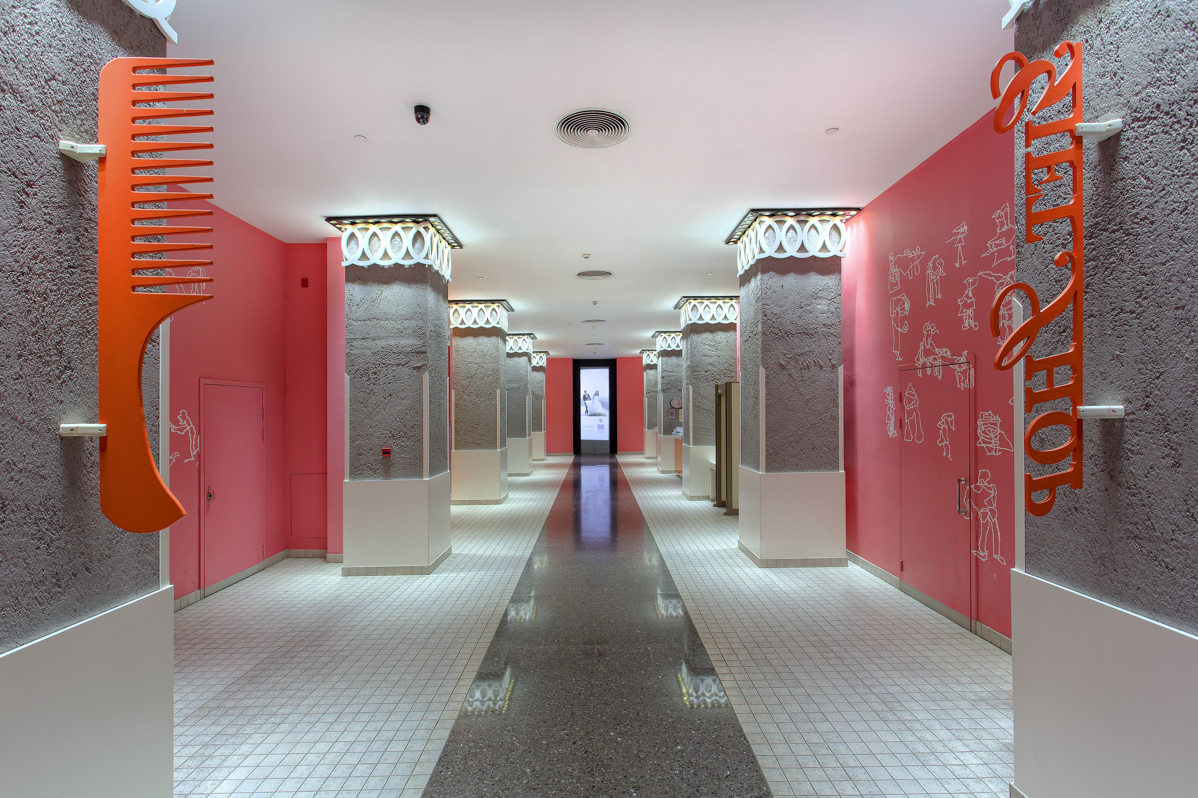 A hotel corridor with a central reflective floor, pink walls, and columns with decorative tops.