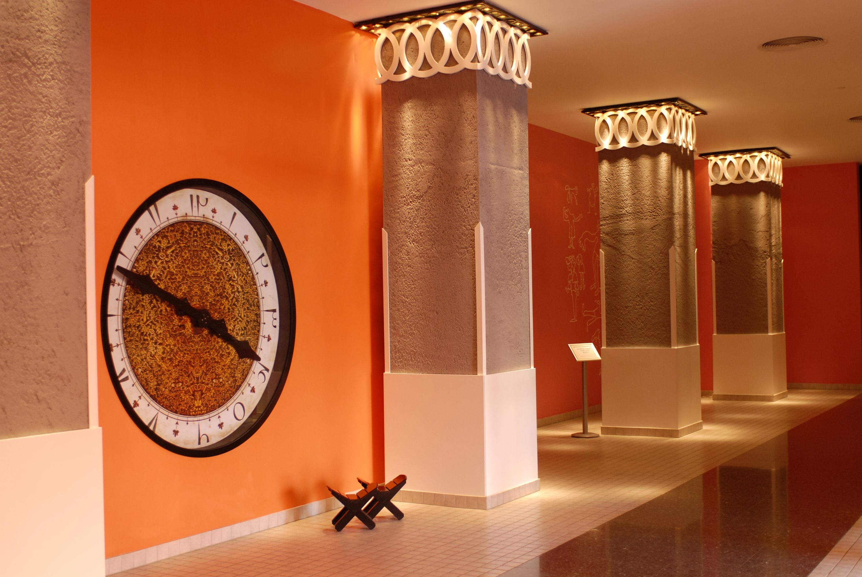 Un pasillo con un reloj de pared grande, columnas de diseños intrincados y una pequeña mesa con una silla.