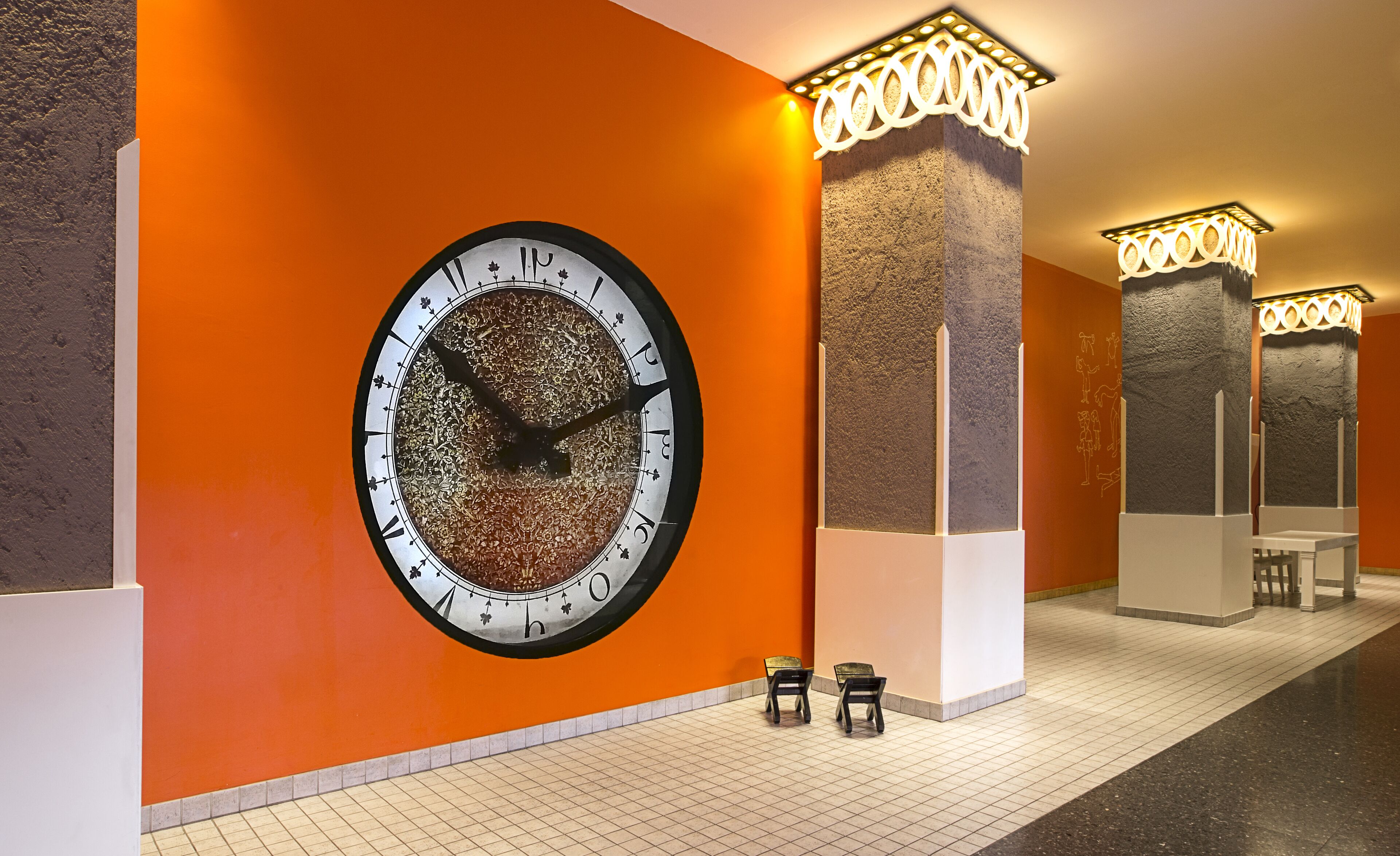 A hallway with an ornate wall clock, tiled flooring, and modern light fixtures.