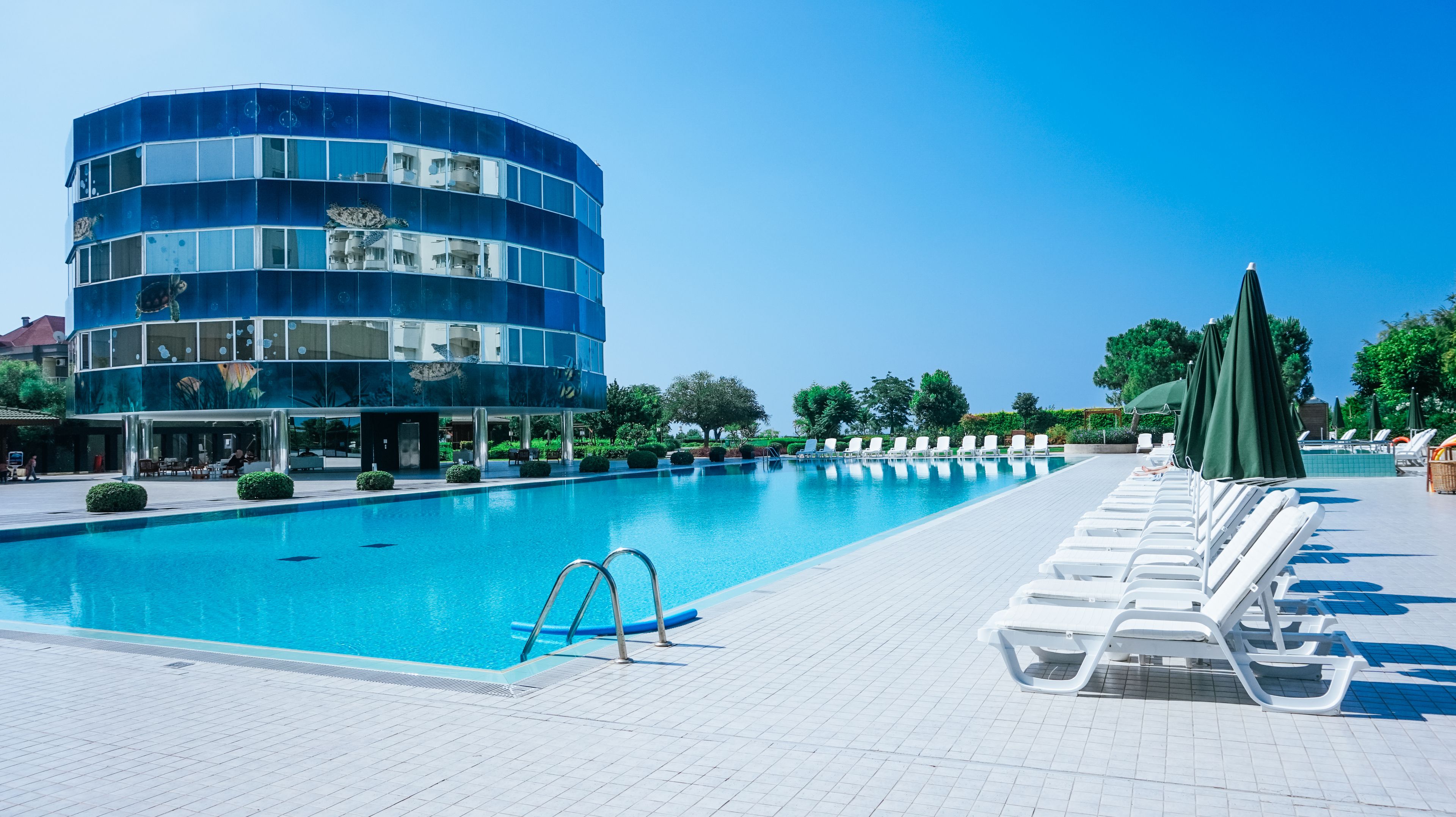 A swimming pool with lounge chairs and umbrellas, surrounded by a building with a blue facade and a clear sky.