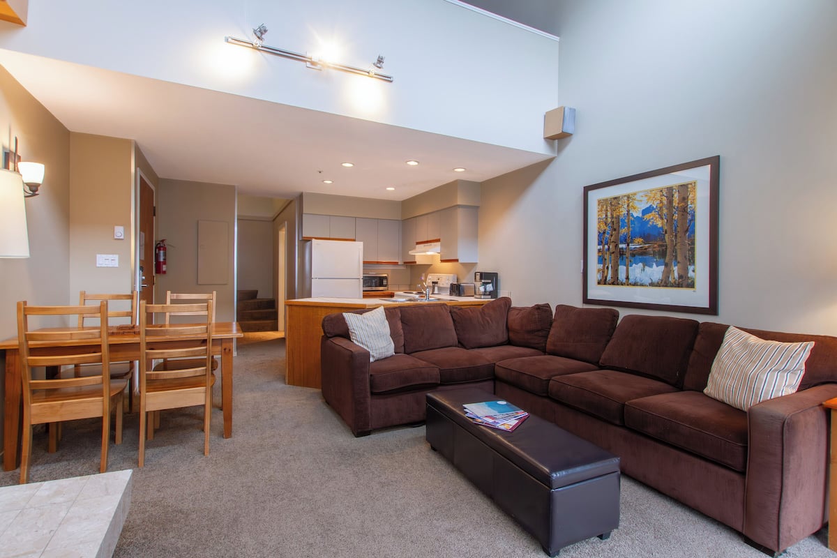 A modern living room with a brown sectional sofa, wooden chairs, a coffee table, and a kitchen in the background.