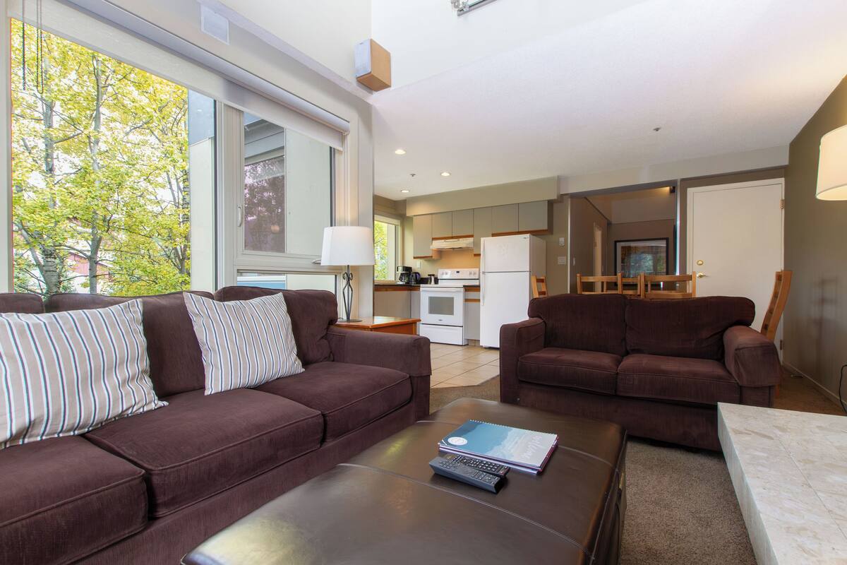 A modern living room with two brown sofas, a coffee table, and a view of a kitchen and dining area.