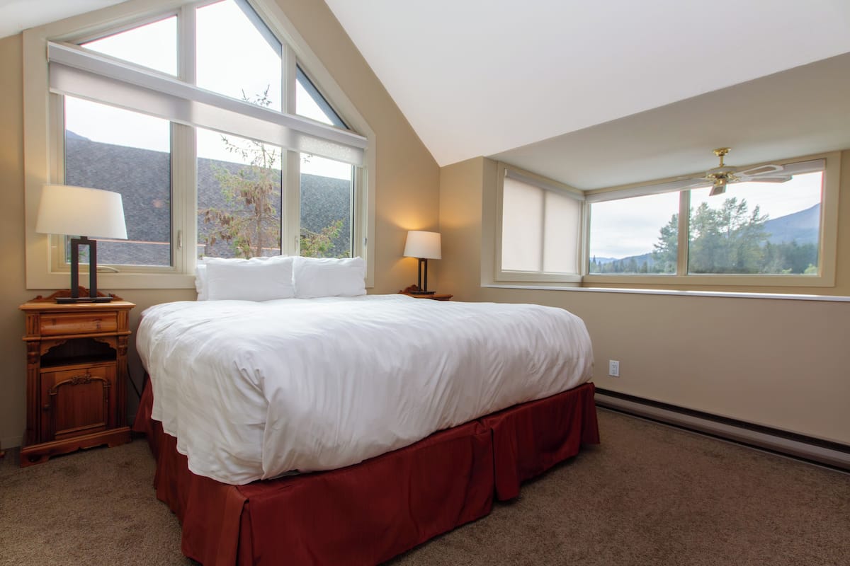 A bedroom with a large bed, two bedside tables, a ceiling fan, and large windows with a mountain view.