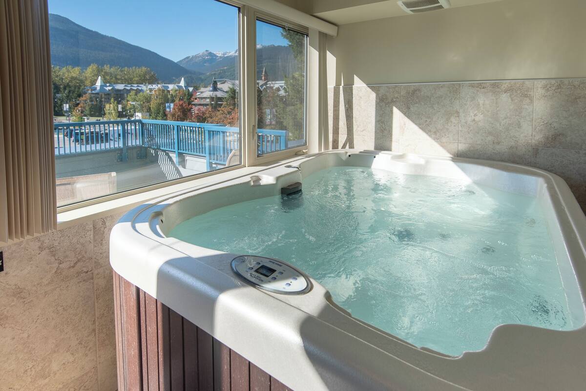 A hot tub with a control panel, a window with a view of mountains and a blue railing outside.