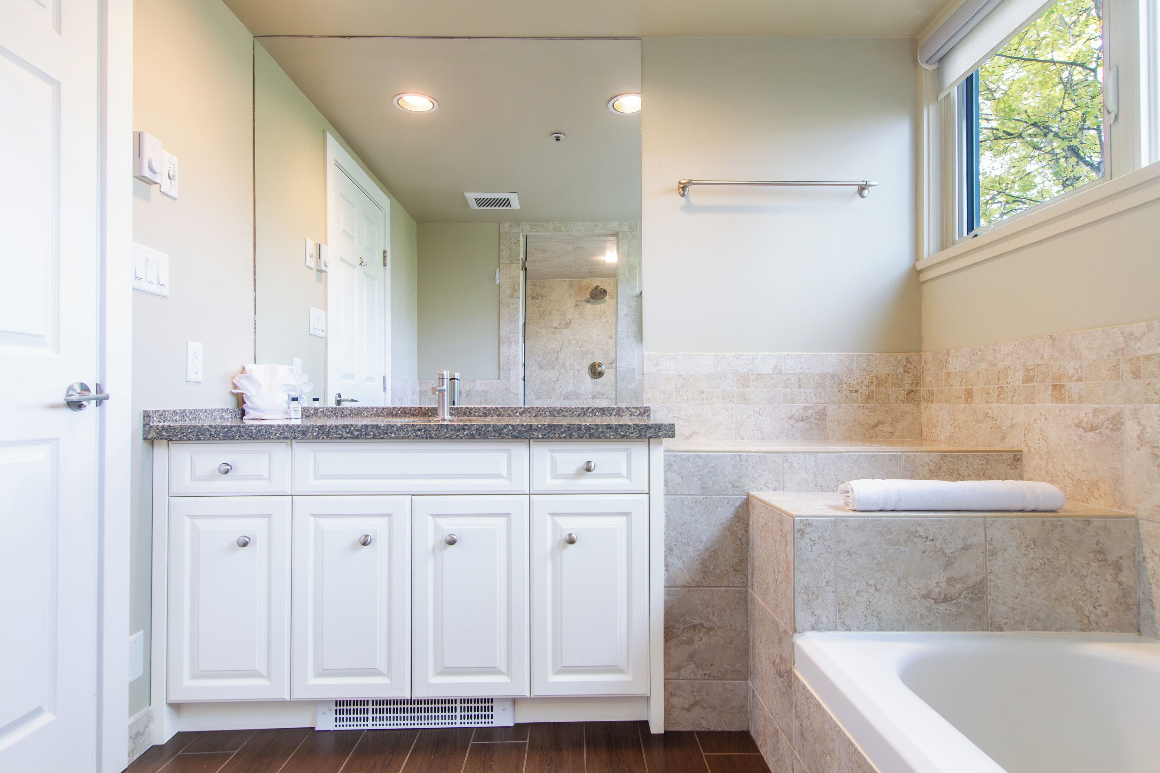 Une salle de bain avec des meubles blancs, un comptoir en granit, une baignoire et une fenêtre donnant sur les arbres.