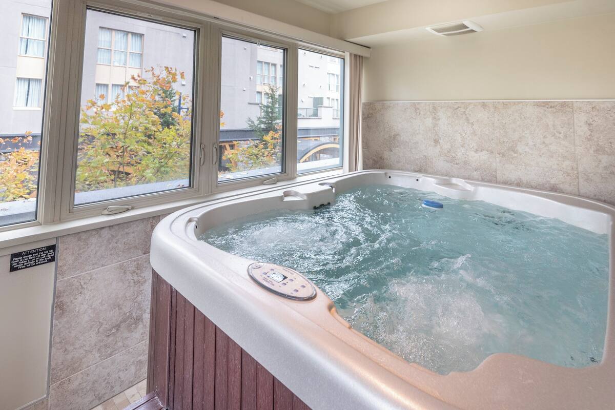 A hot tub with a control panel, a window with a view of buildings and trees, and tiled walls.