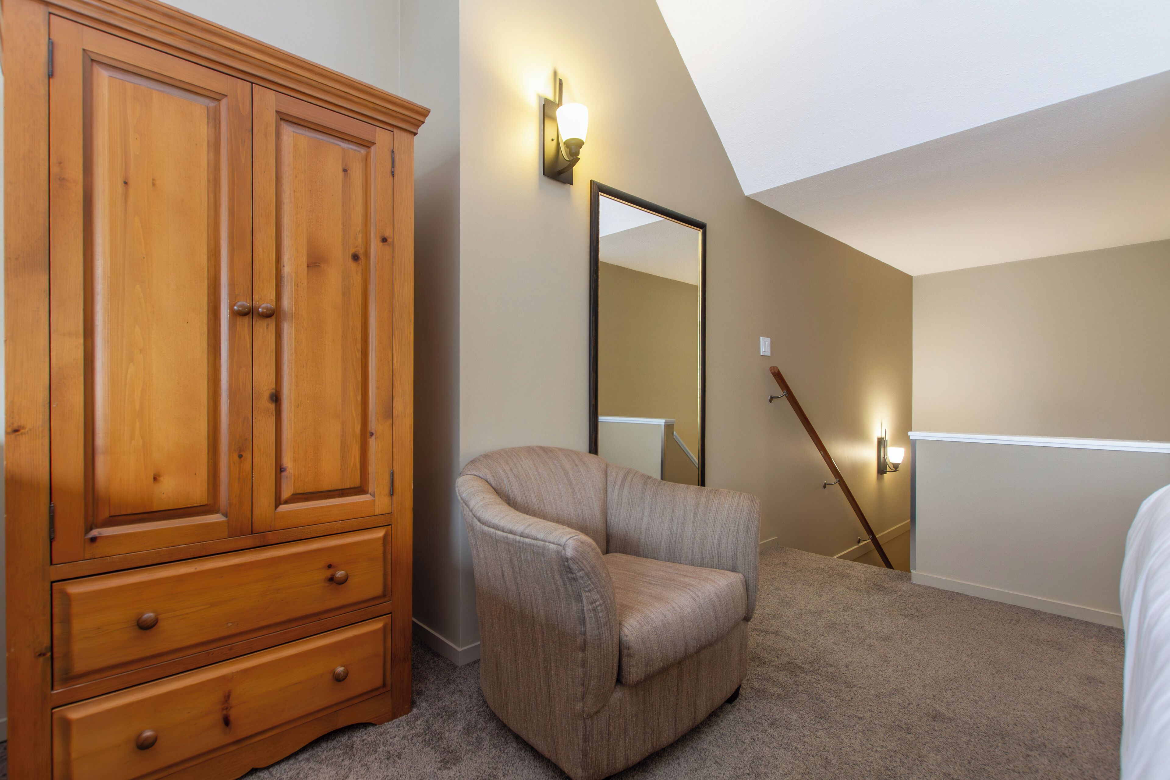 A bedroom with a wooden wardrobe, a beige armchair, a full-length mirror, and a sloped ceiling.