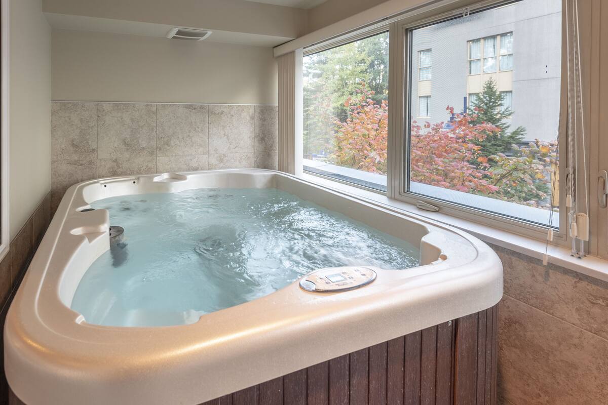 A hot tub in a room with a large window, a tiled wall, and a view of trees and buildings outside.