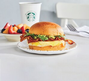 Un sándwich de desayuno en un plato, con una taza y un bol de fruta al fondo.