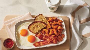 Un piatto per la colazione con uova, pane tostato, pancetta e patate arrosto su un tavolo di legno.