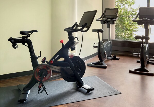 Una bicicleta estática con pantalla digital y un tablet, ubicada en una habitación con ventanas amplias y otra bicicleta estática al fondo.