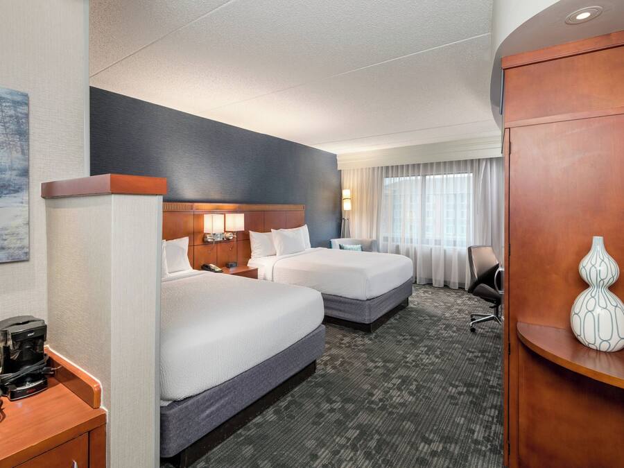 A hotel room with two beds, a desk with a coffee maker, a lamp, and a vase on a wooden sideboard.