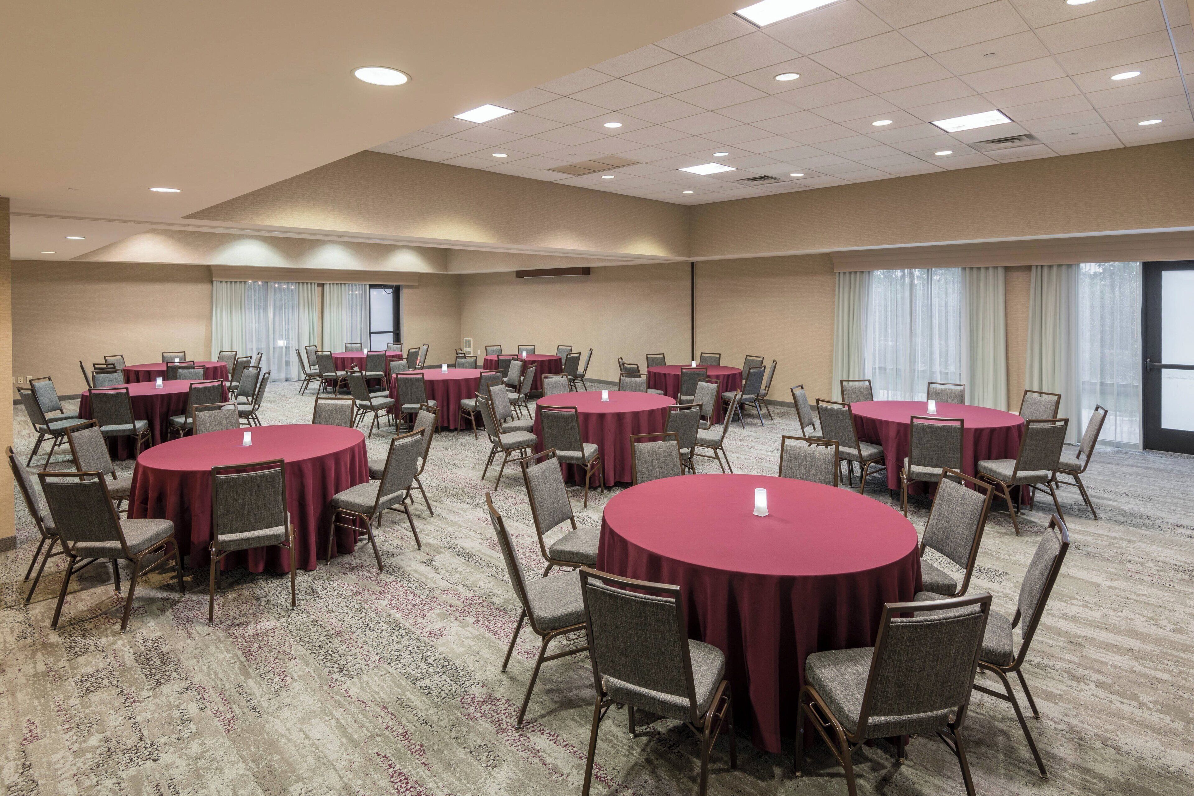 A conference room with multiple round tables and chairs, carpeted flooring, and large windows with curtains.