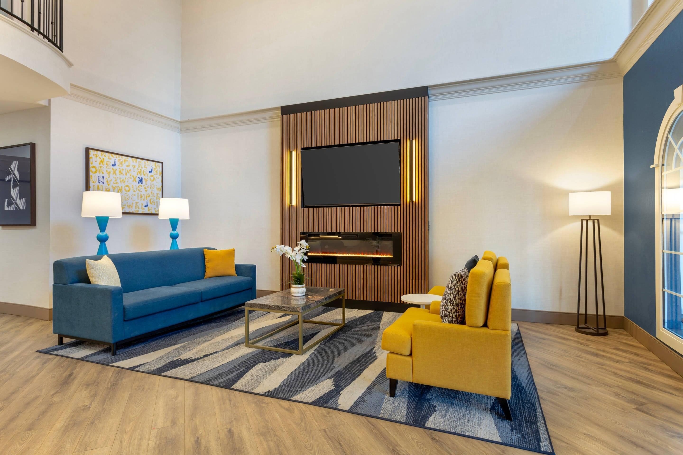 A modern living room with a blue sofa, yellow armchair, and a coffee table. A flat-screen TV is mounted on the wall above a wooden panel.