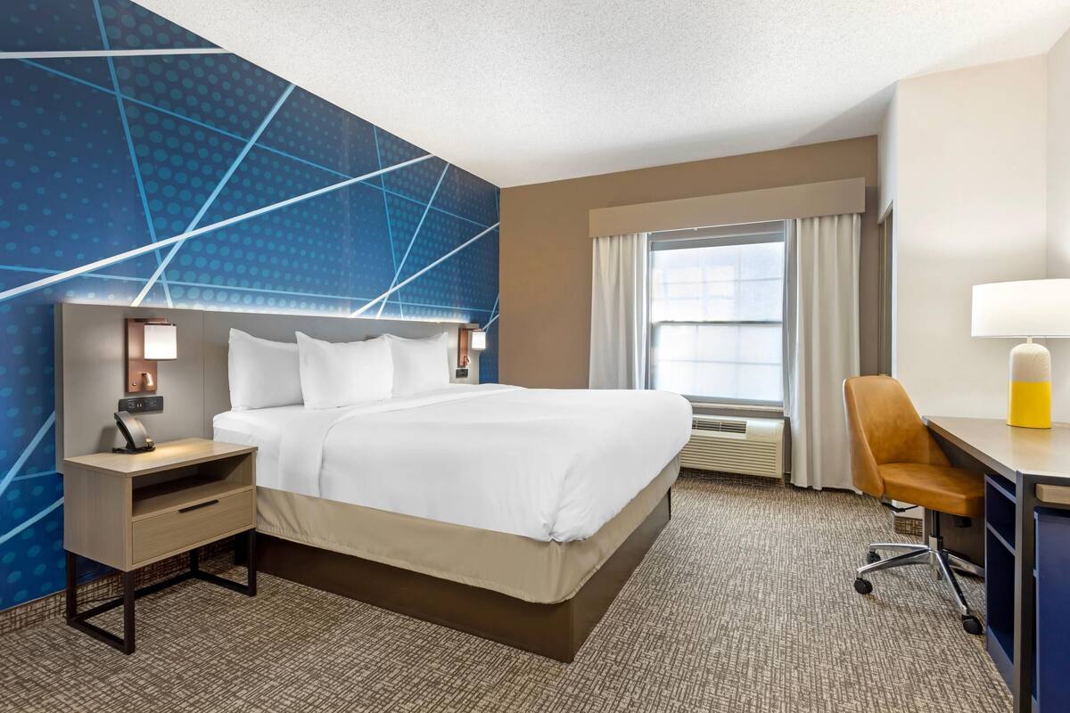 Ein Hotelzimmer mit einem großen Bett, einem Schreibtisch mit Stuhl, einem Nachttisch, einer Lampe und einem Fenster mit Vorhängen.