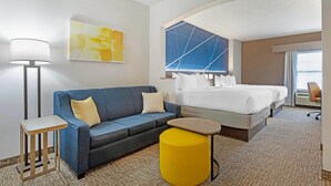Habitación de hotel con un sofá azul, una mesita pequeña, un reposapiés amarillo, una cama con ropa blanca y un ventanal con vistas geométricas.
