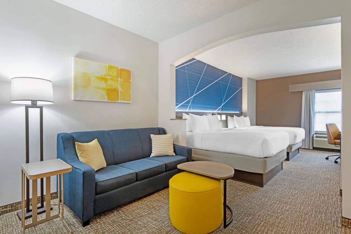 Ein Hotelzimmer mit blauem Sofa, einem kleinen Tisch, einem gelben Hocker, einem Bett mit weißen Bettwäsche und einem großen Fenster mit geometrischer Aussicht.