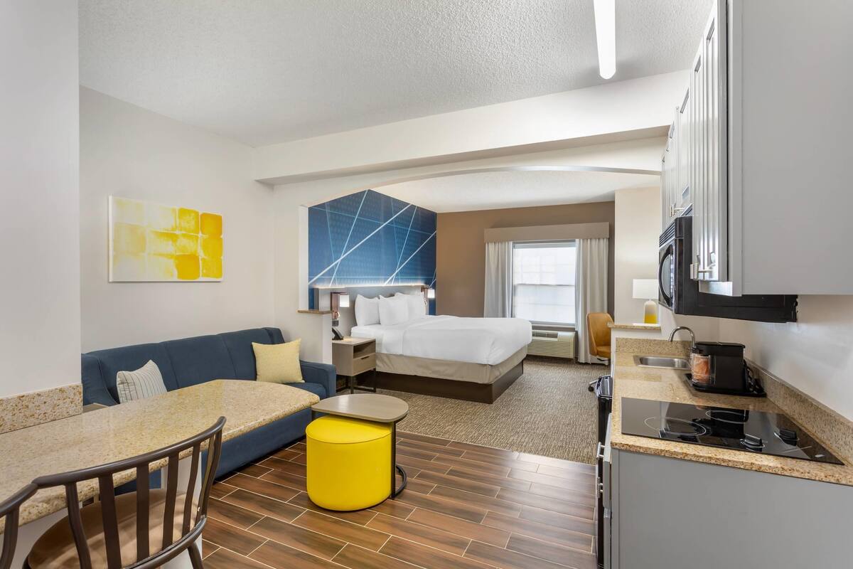 Ein Hotelzimmer mit Küchenzeile, einem Bett, einem Sofa, einem Esstisch und einem gelben Hocker.