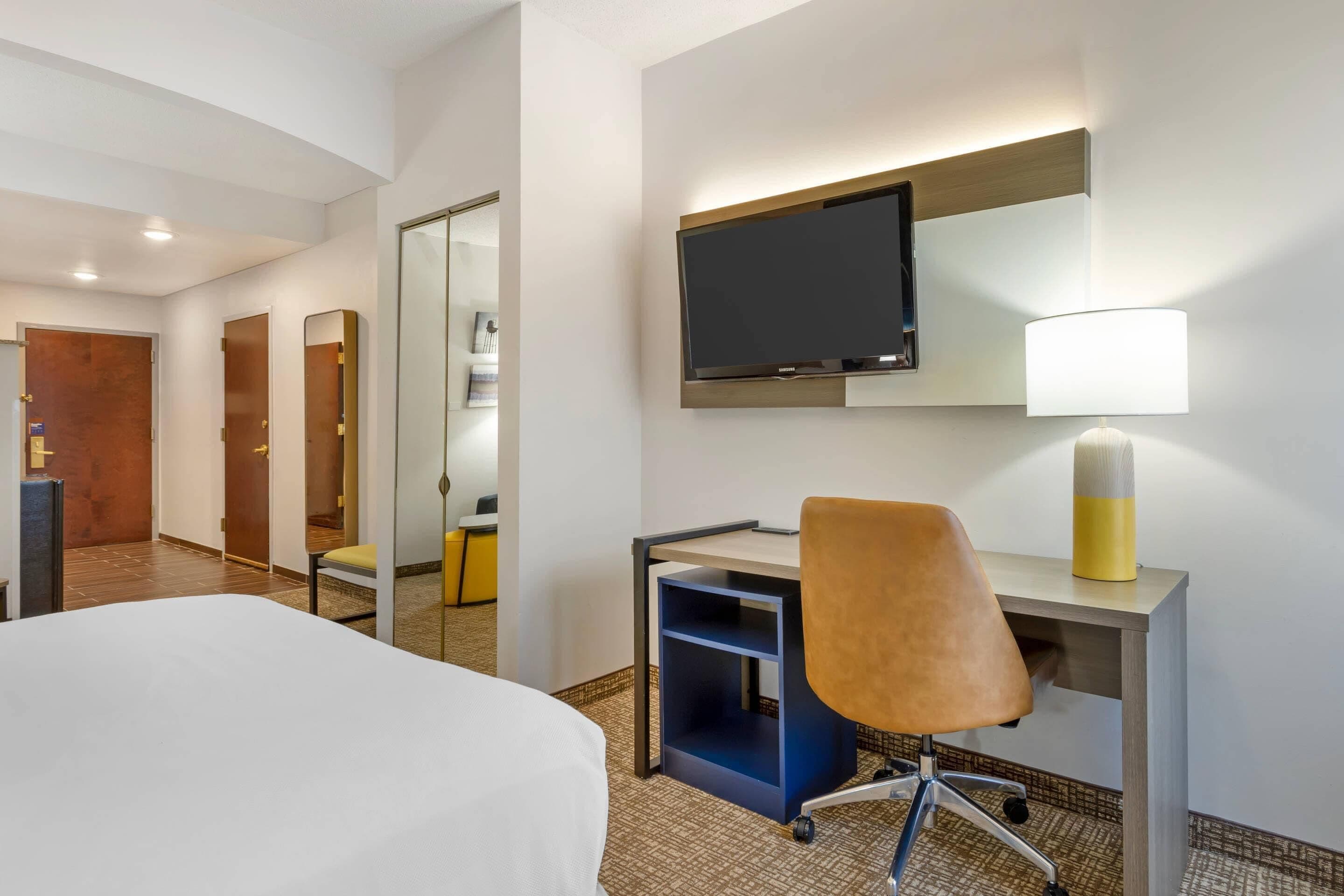 Una habitación de hotel con cama, escritorio, silla, televisión, lámpara y espejo.