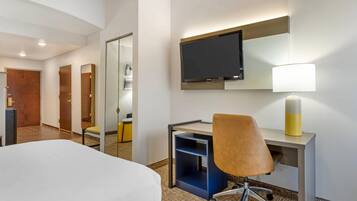 Una habitación de hotel con cama, escritorio, silla, televisión, lámpara y espejo.