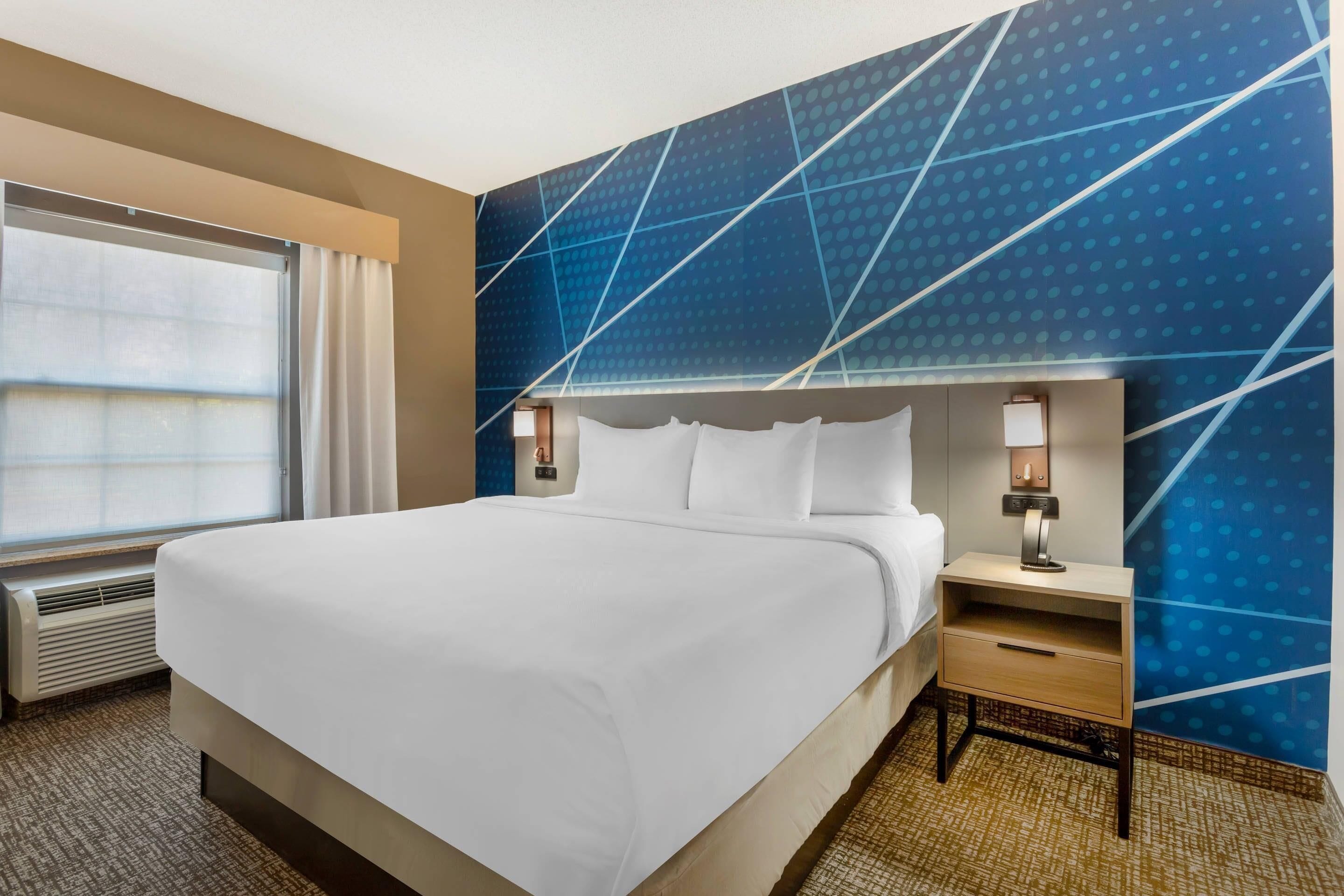 Ein Hotelzimmer mit einem großen Bett, einem Nachttisch mit Lampe, einem Fenster mit Vorhängen und einer Wand mit geometrischem Muster.