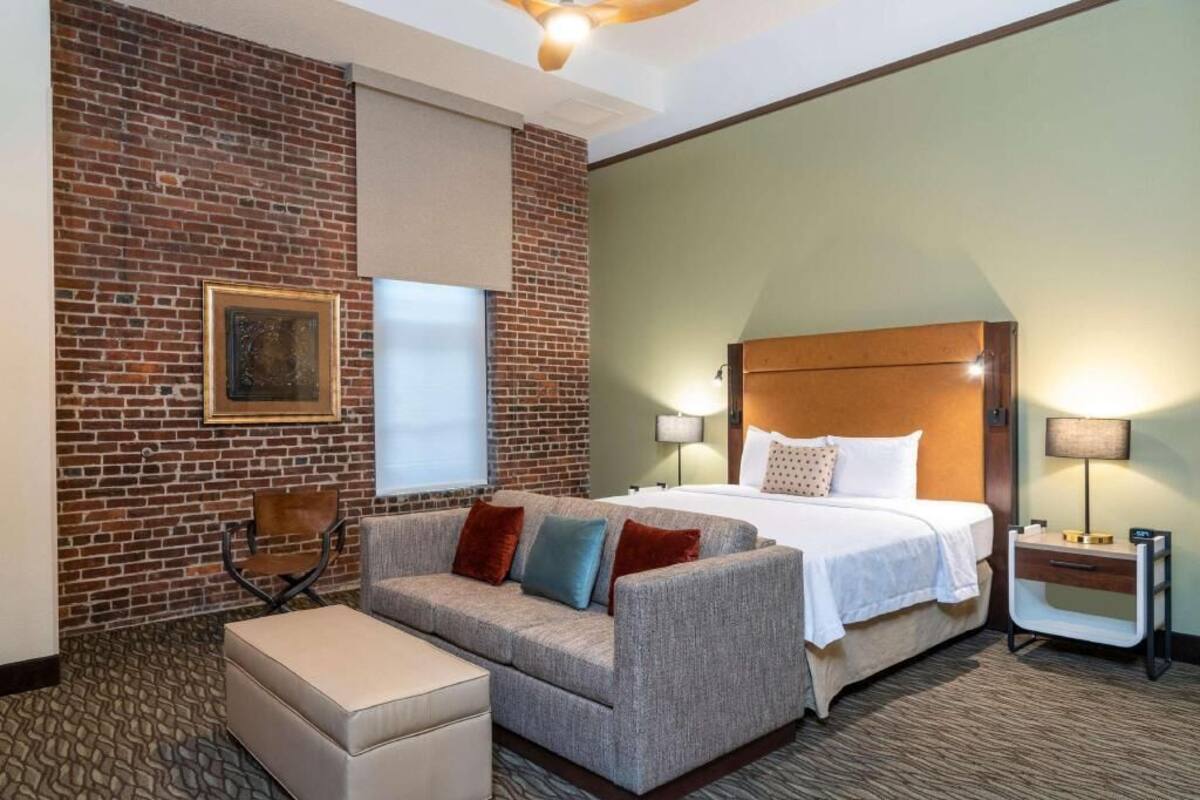 Habitación de hotel con pared de ladrillo, cama con cabecera de madera, sofá con cojines, una mesita y una ventana con persianas.