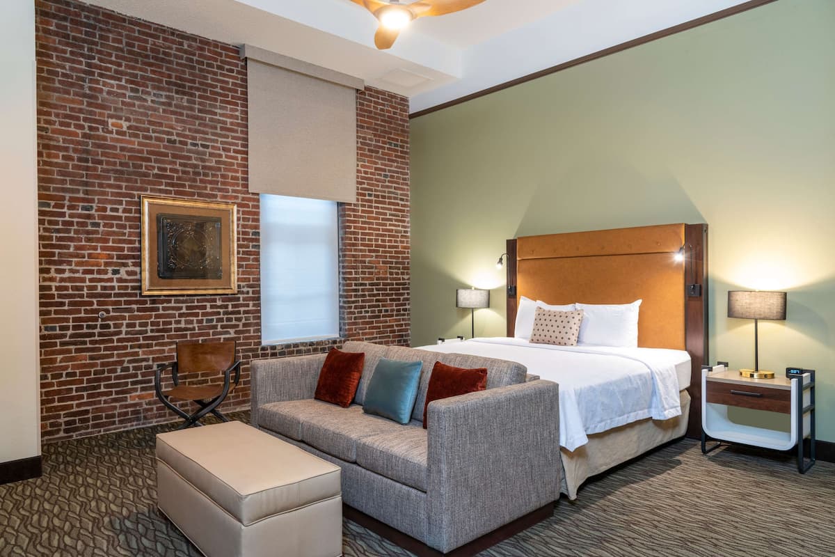 Ein Hotelzimmer mit Backsteinwand, einem großen Bett mit weißen Leinen, einem grauen Sofa mit vielen Kissen, einem kleinen Tisch und einem Stuhl.