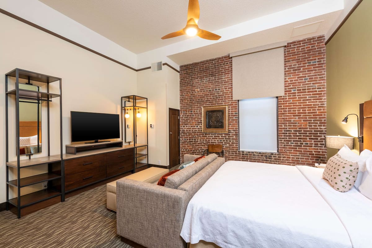 Ein modernes Hotelzimmer mit Ziegelakzentwand, einem großen Bett, einem Flachbildfernseher, einer Couch und einem Deckenventilator.