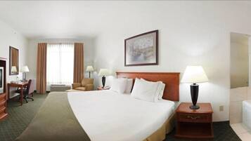 A hotel room with a large bed, a desk with a chair, a small table with a lamp, and a large window with curtains.