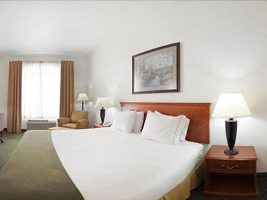 A hotel room with a large bed, a desk with a chair, a small table with a lamp, and a large window with curtains.