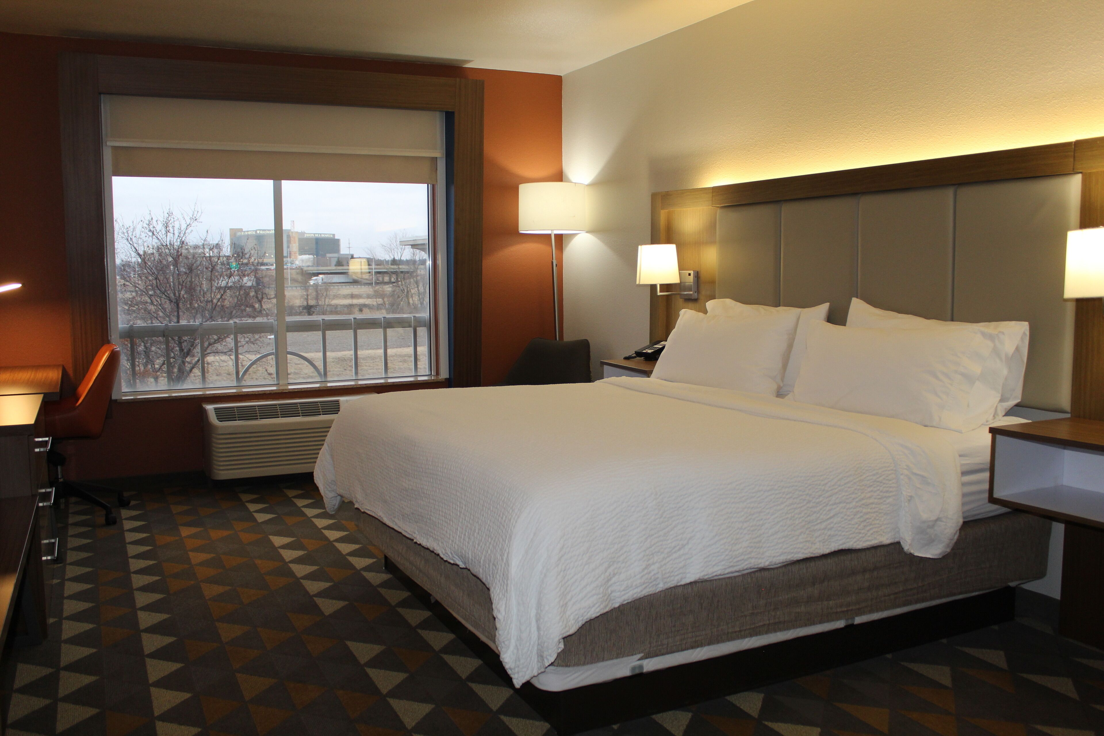 Habitación de hotel con una cama grande, un escritorio, una silla, un ventanal con vistas a edificios y un suelo estampado.