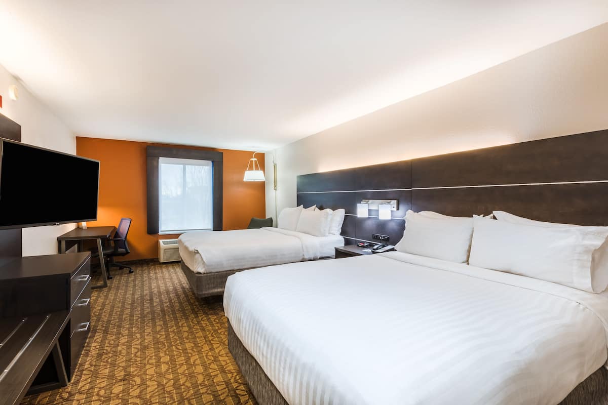 Ein Hotelzimmer mit zwei Betten, einem Schreibtisch, einem Stuhl, einem Flachbildfernseher und einem Fenster mit Jalousien.