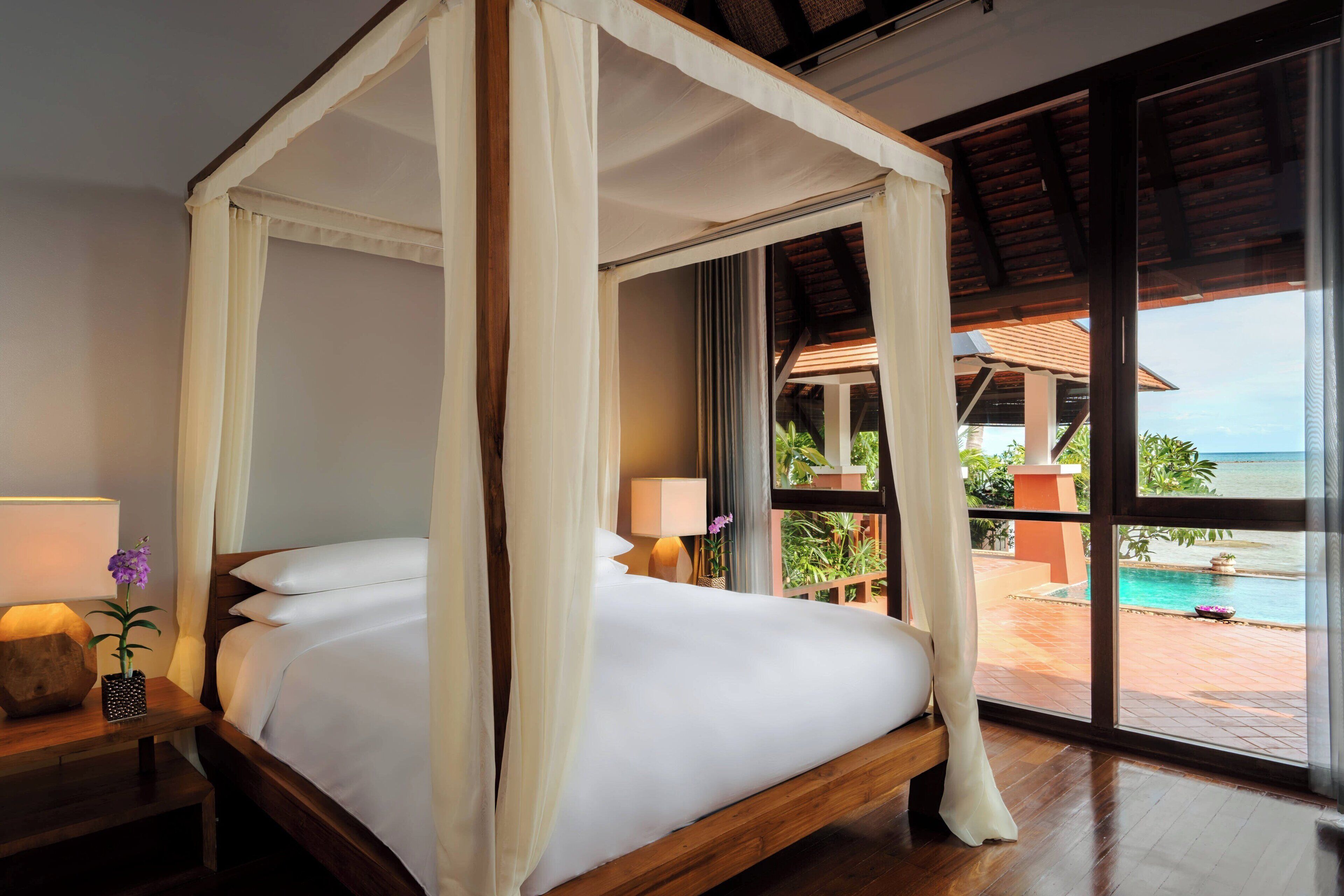 A four-poster bed with white linens, a wooden nightstand with a lamp, and a view of a pool and greenery through large windows.