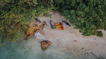 An aerial view of a beach with kayaks and a surfboard, surrounded by dense foliage.