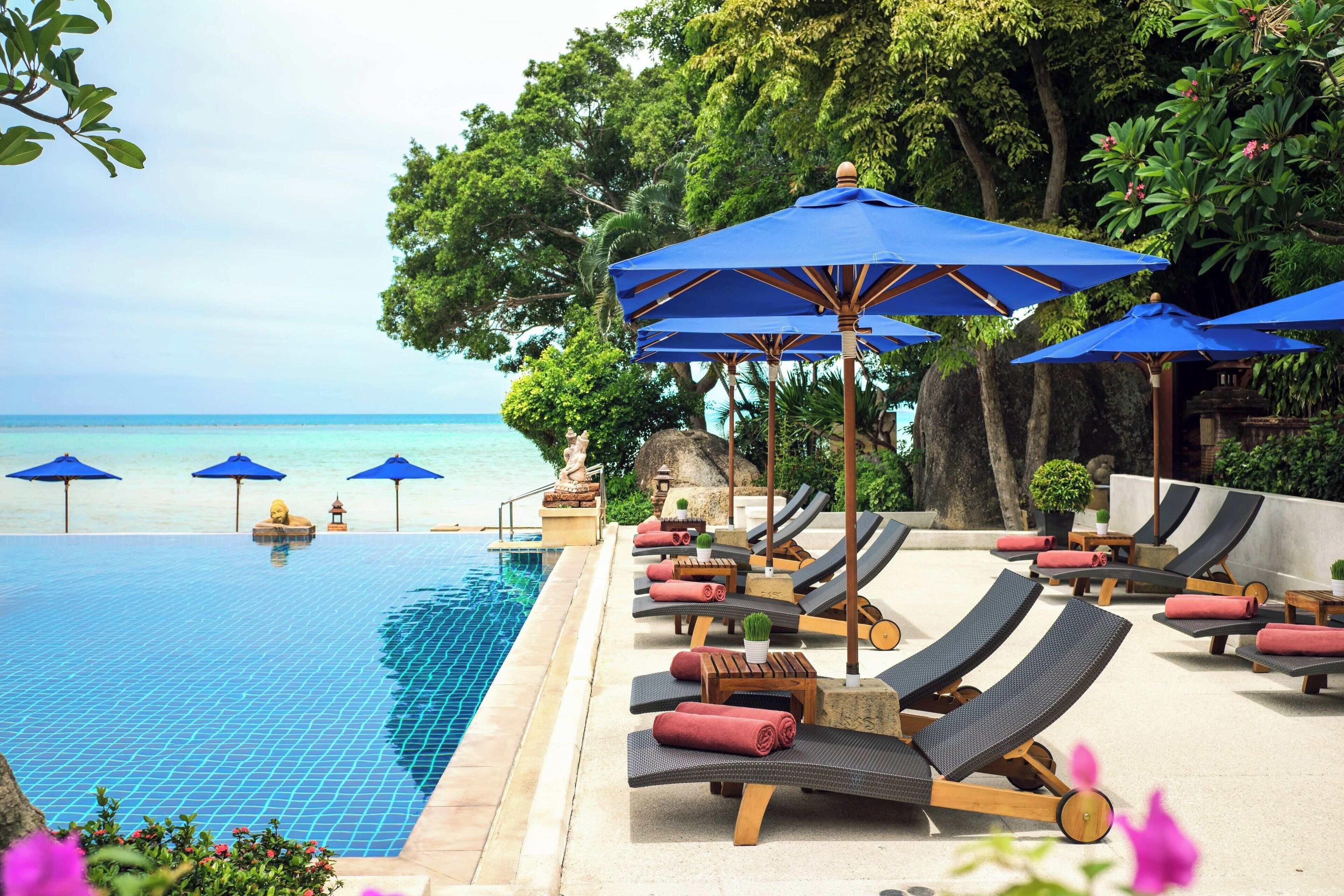 A poolside area with lounge chairs and blue umbrellas, overlooking a clear blue sea.