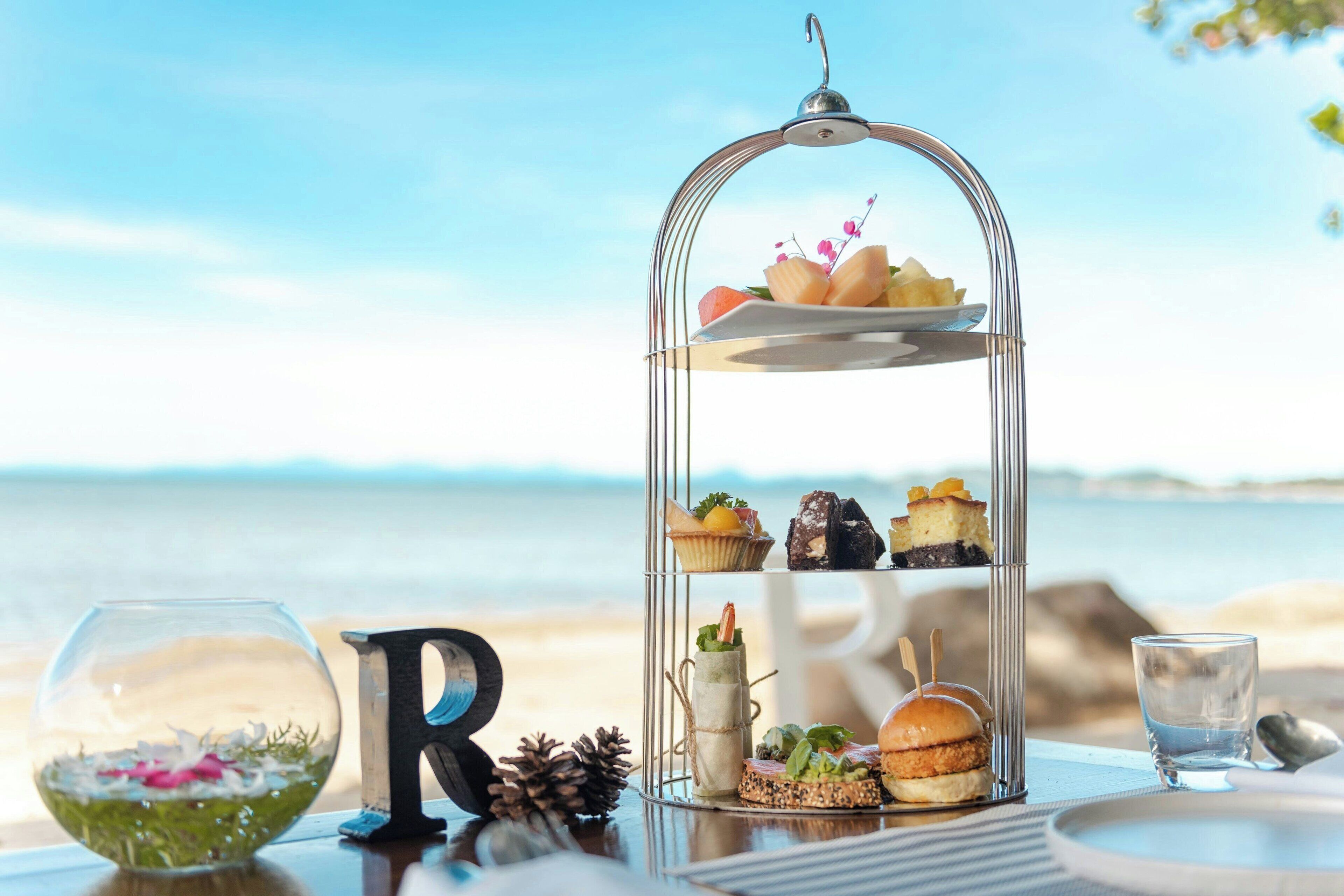 Una mesa frente al mar con un soporte de varios niveles para postres, un vaso con un arreglo floral y una decoración con la letra 'R'.