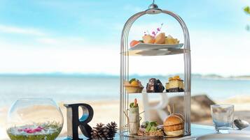 Una mesa frente al mar con un soporte de varios niveles para postres, un vaso con un arreglo floral y una decoración con la letra 'R'.