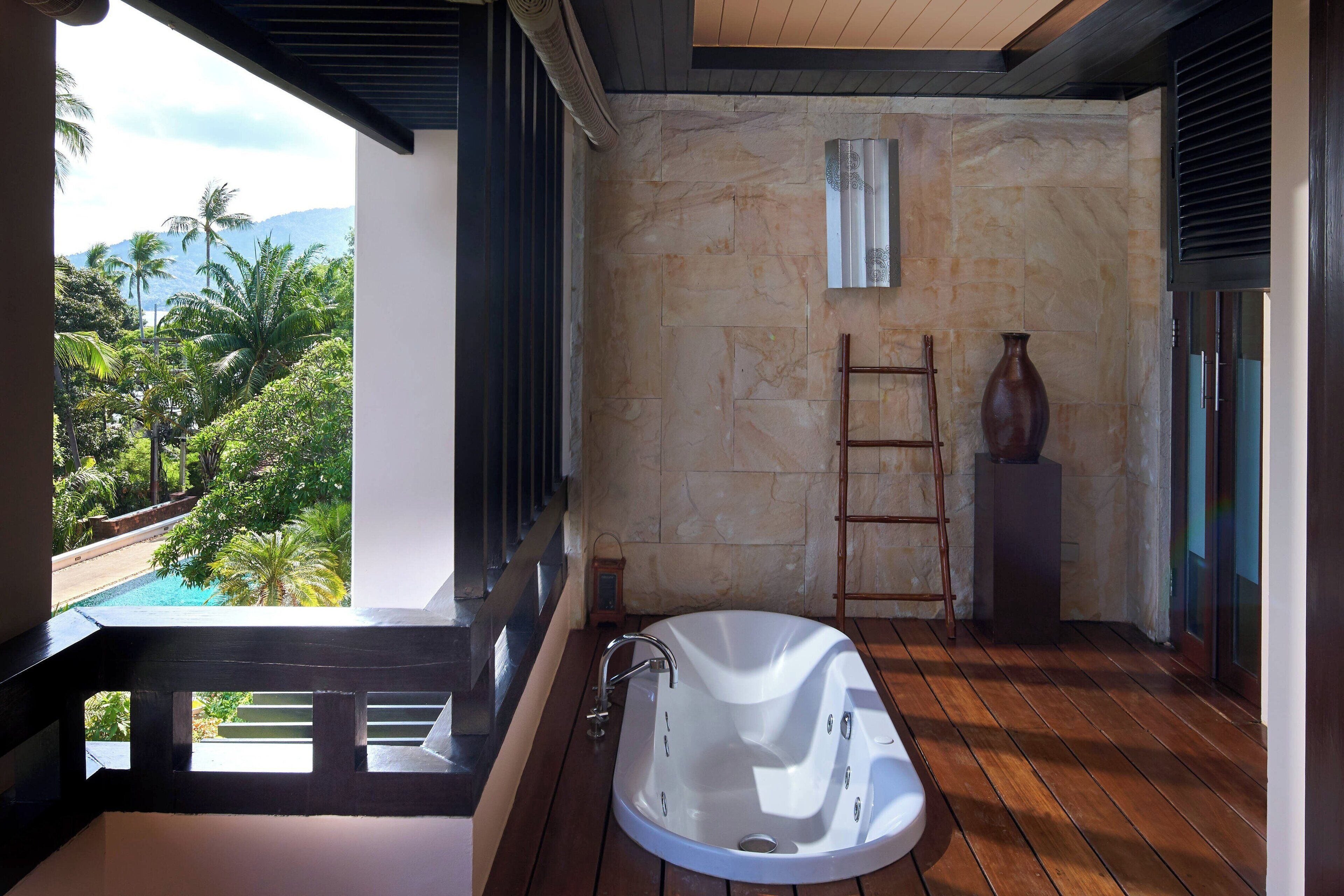 Ein modernes Badezimmer mit freistehender Badewanne, einer Steinwand und Blick auf ein Schwimmbecken und Palmen.