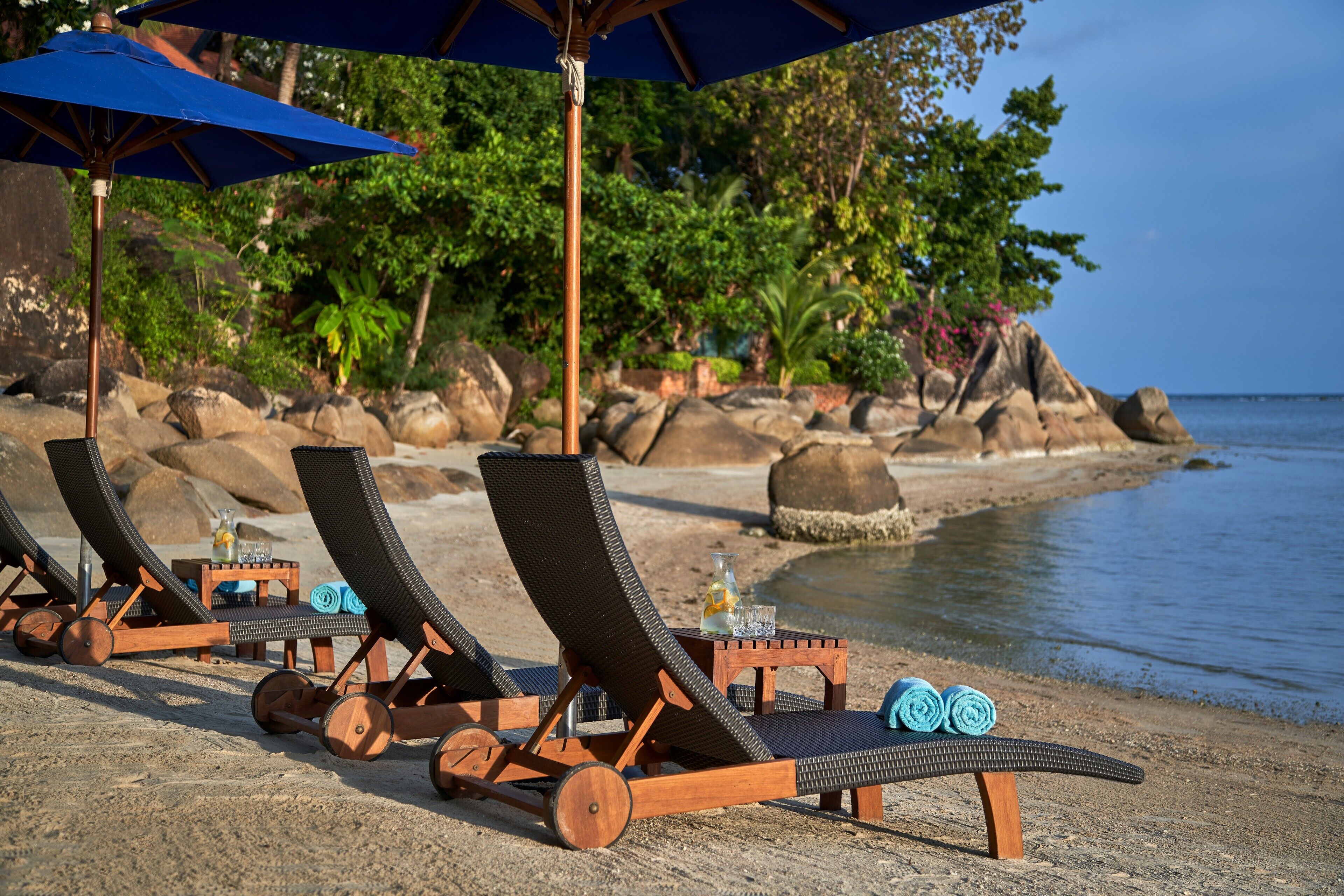 Un espace de détente aménagé sur la plage, avec des chaises longues en bois et des parasols bleus, une petite table avec une bouteille et un verre, et une vue sur l’océan et la végétation en arrière-plan.